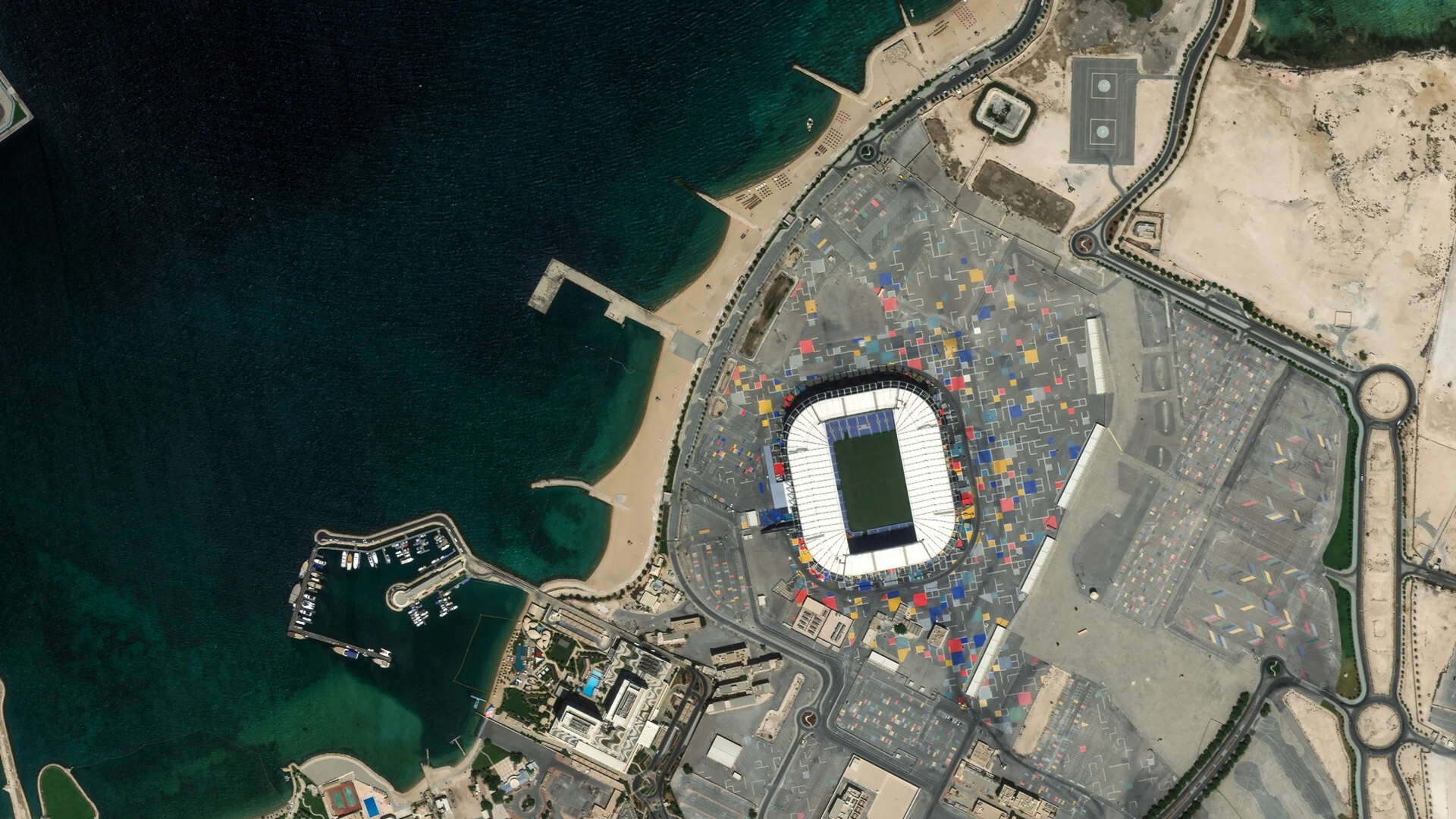 Pelican image of a waterfront stadium built from stacked shipping containers, beside a marina and a sweep of engineered beach