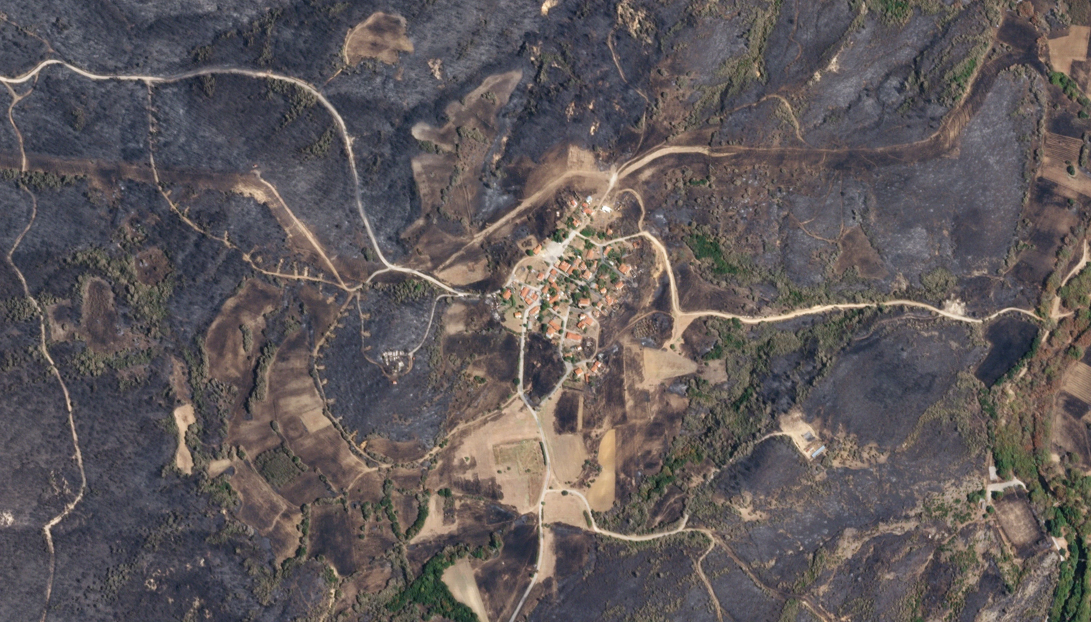 SkySat image of a hillside village left standing inside a large burn scar, its orange roofs surrounded by scorched ground and pale dirt tracks